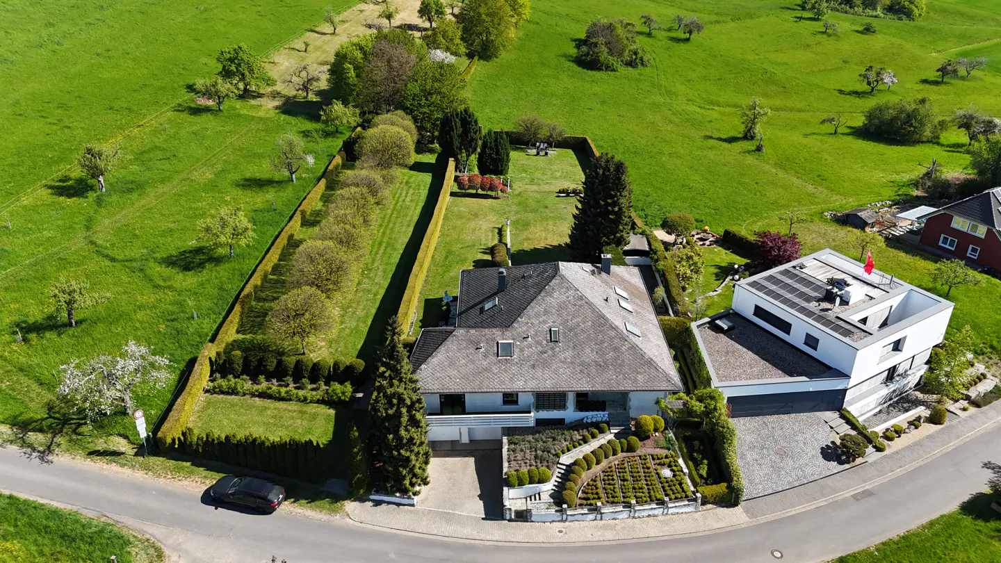 Aerial view of a gray-roofed house with a green lawn, surrounded by tall hedges, next to a modern white house with solar panels.
