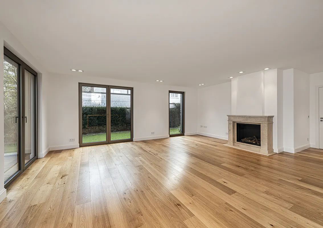 Bright, empty living room with hardwood floors, white walls, and a stone fireplace. Glass doors lead to a green yard.