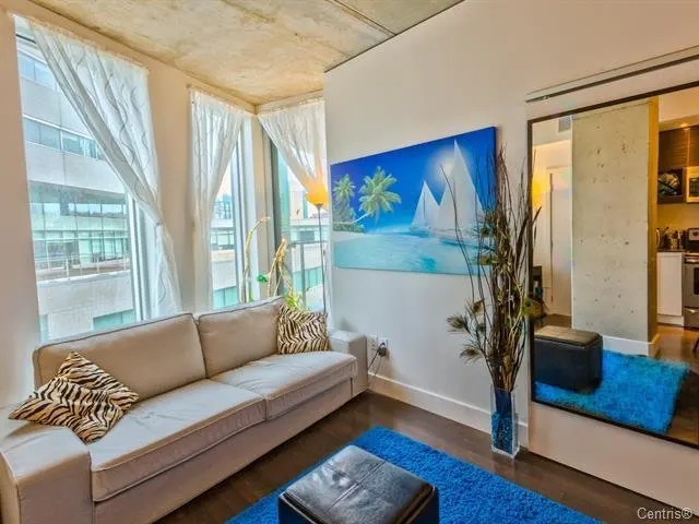Living room with a beige sofa, zebra-print pillows, and a blue ocean painting. White curtains filter light from large windows. A blue rug adds color.