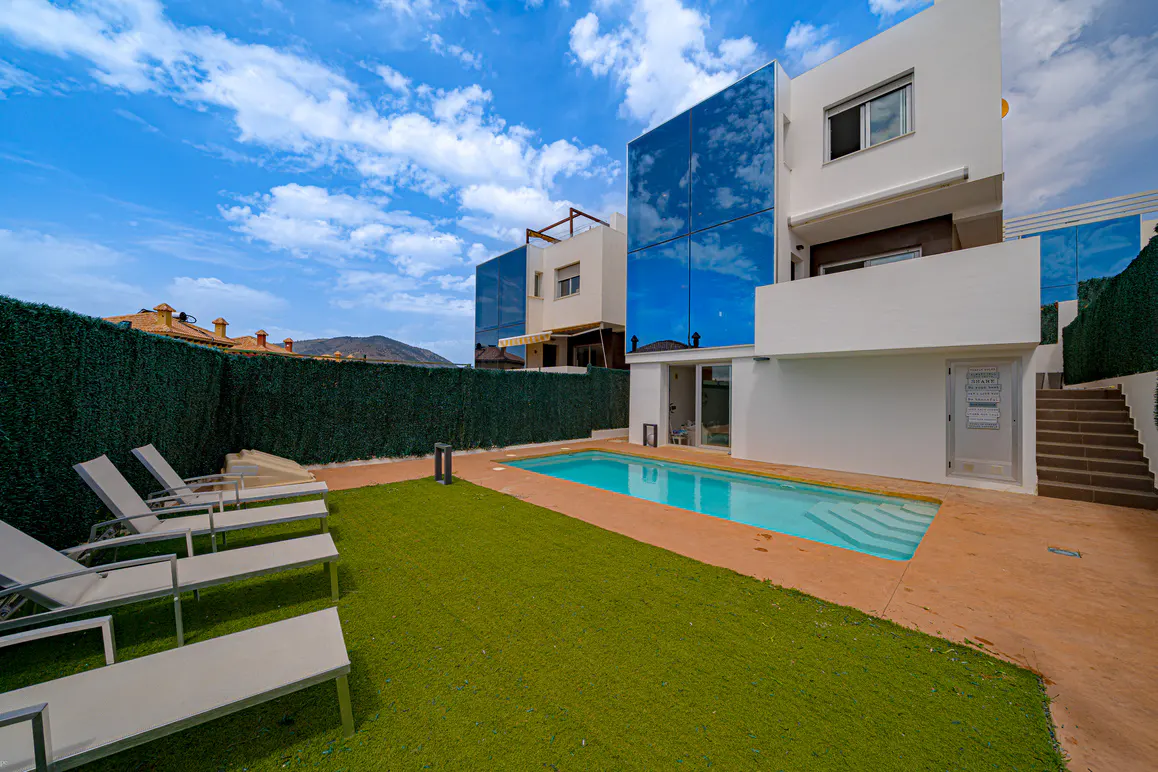 Modern white house with a pool, green lawn, and lounge chairs under a blue sky with clouds. The house has blue reflective glass.