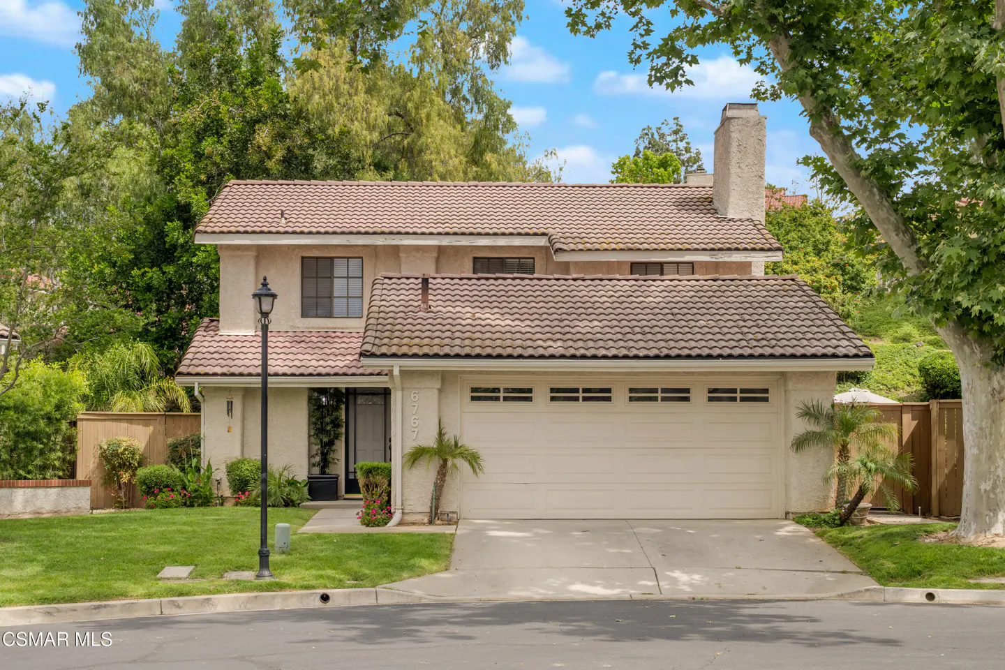 Two-story beige house with a brown tile roof, a two-car garage, and a green front lawn under a blue sky.