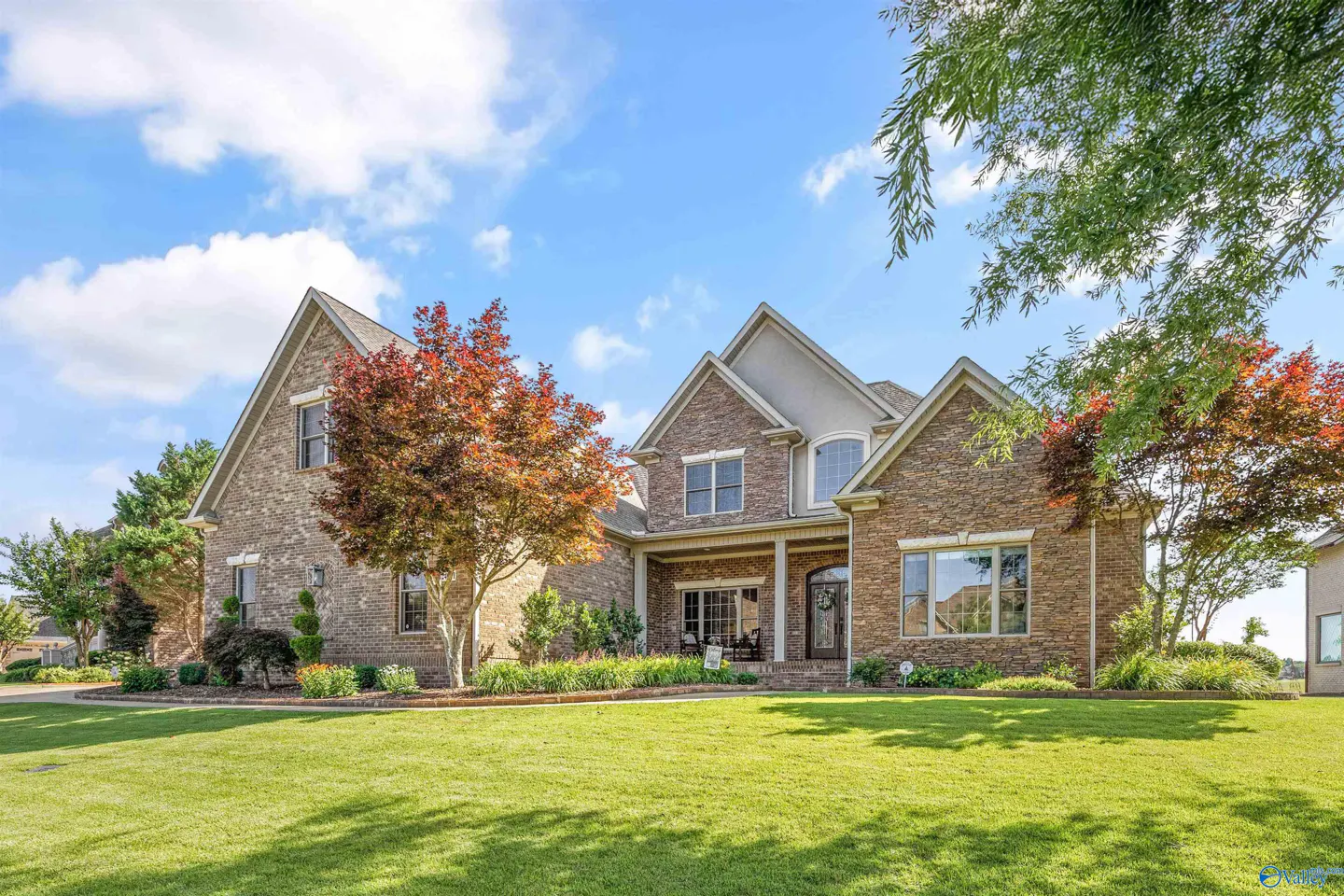 Two-story brick house with a gabled roof, manicured lawn, and vibrant red-leafed trees under a bright blue sky.
