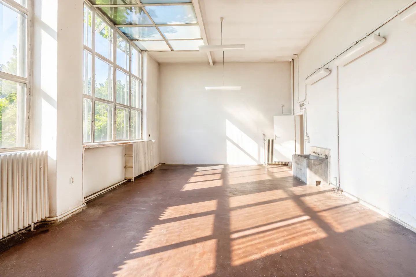 Bright, empty industrial-style room with large windows, a skylight, white walls, brown floors, and a utility sink.