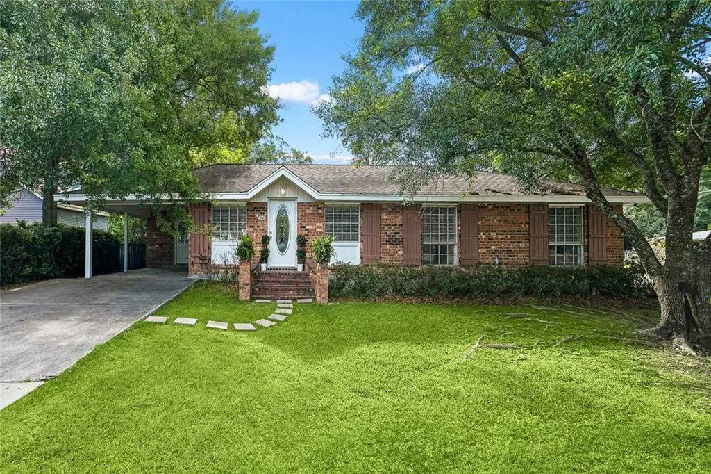 A single-story brick house with brown shutters and a white front door. A stone path leads to the door, and a carport is on the left.