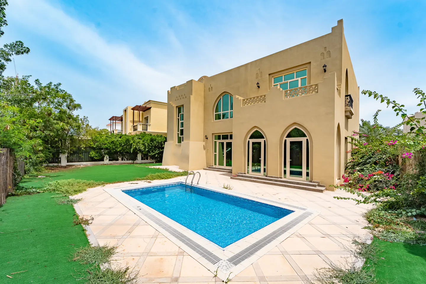 Beige two-story house with arched windows and a blue swimming pool in the backyard under a blue sky.