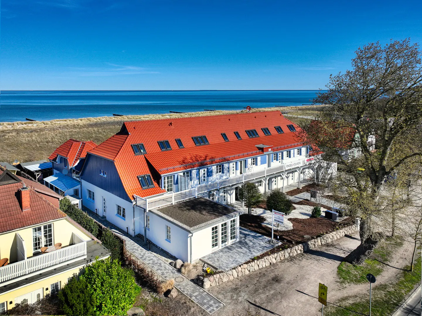 Aerial view of a multi-story building with a red roof, white facade, and blue shutters near a sandy beach and blue ocean under a clear sky.