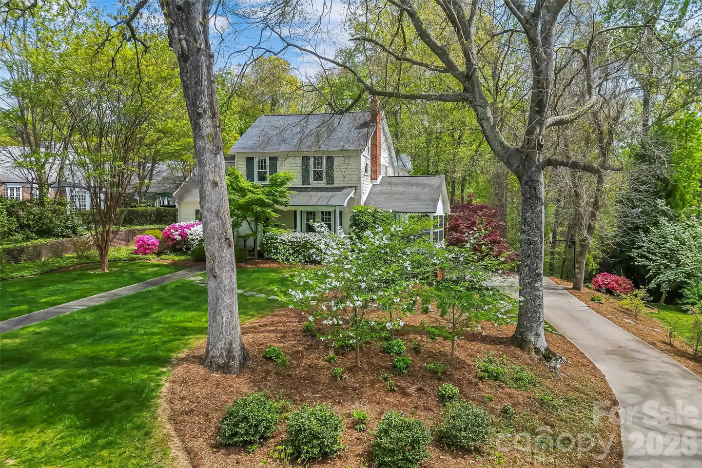 Two-story house with a gray roof, light yellow siding, and green shutters, surrounded by trees and flowering bushes.
