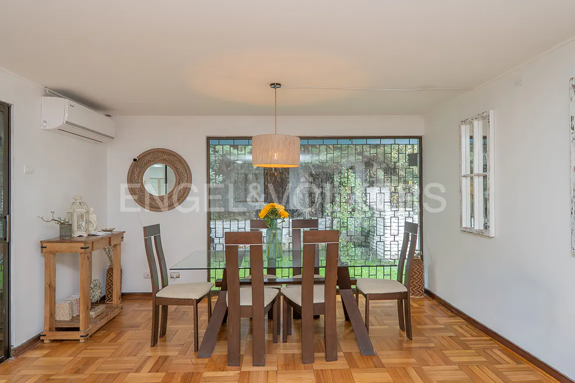Bright dining room with wood floors, a glass-topped table, and wooden chairs. A large window overlooks greenery.