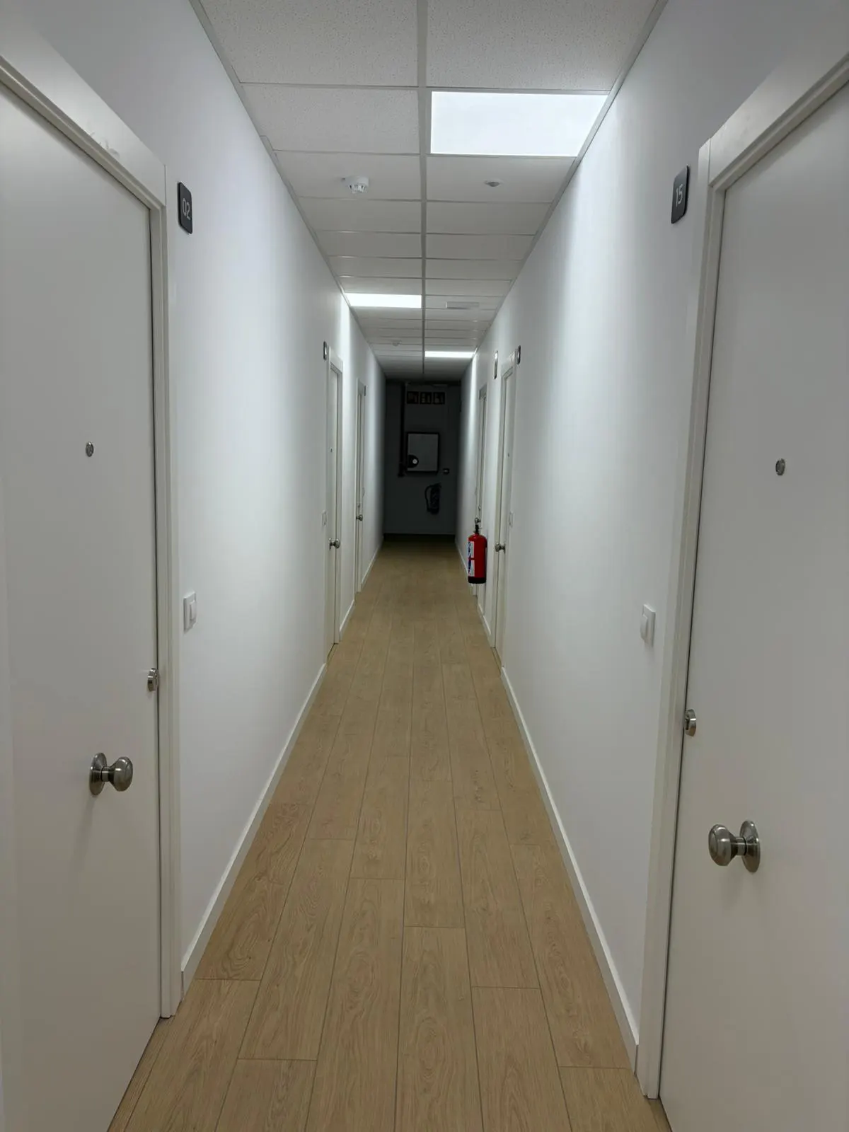 A long, brightly lit hallway with white walls, light wood floors, and white doors on either side. A red fire extinguisher is visible down the hall.