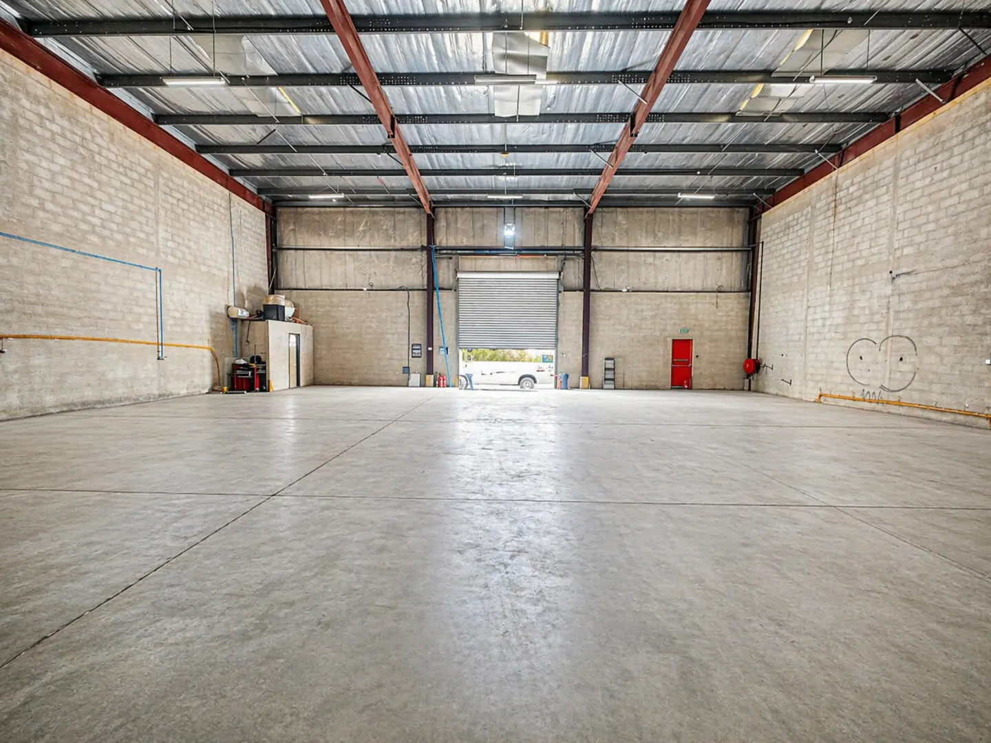 A large, empty warehouse with concrete floors and cinder block walls. A partially open garage door reveals a vehicle outside.