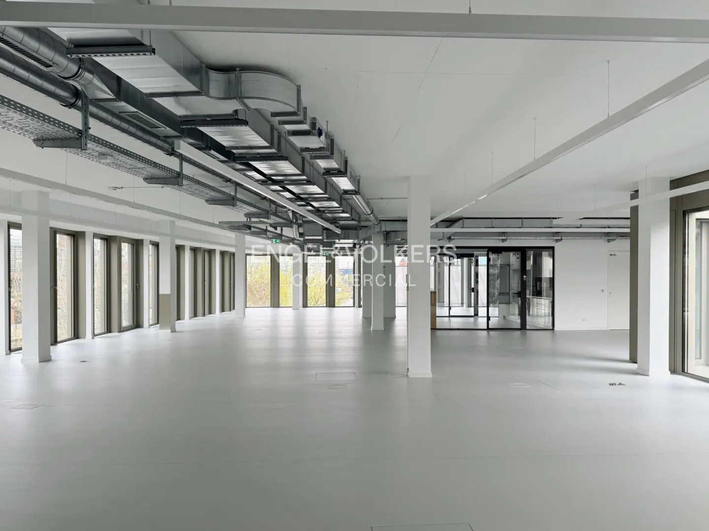 Bright, empty office space with white walls, gray floors, and large windows. Overhead ductwork and support columns are visible.