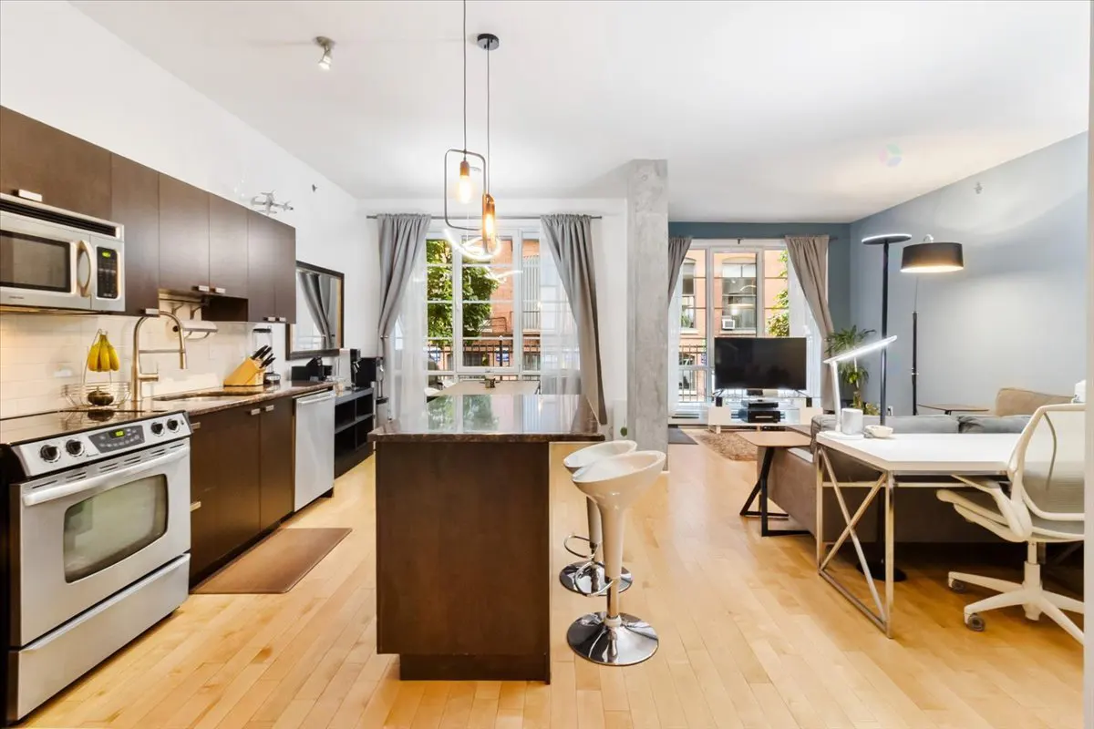 Open-concept apartment with a kitchen featuring dark cabinets, stainless steel appliances, and an island with white stools. Living area with a TV and desk.