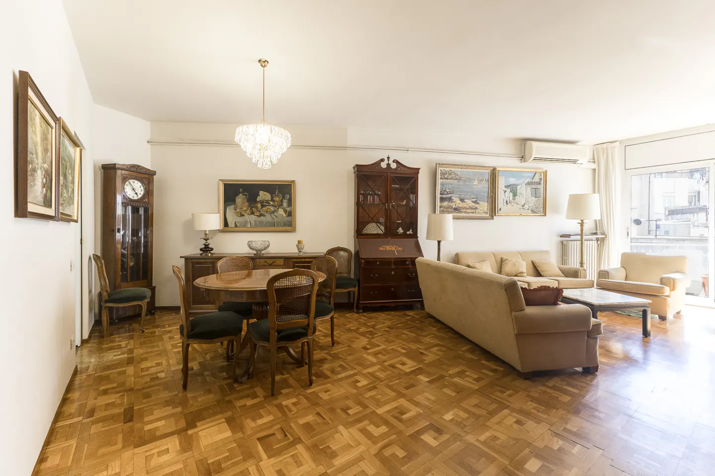 Bright living and dining room with parquet floors, tan sofas, and dark wood furniture. A chandelier hangs above a round dining table.