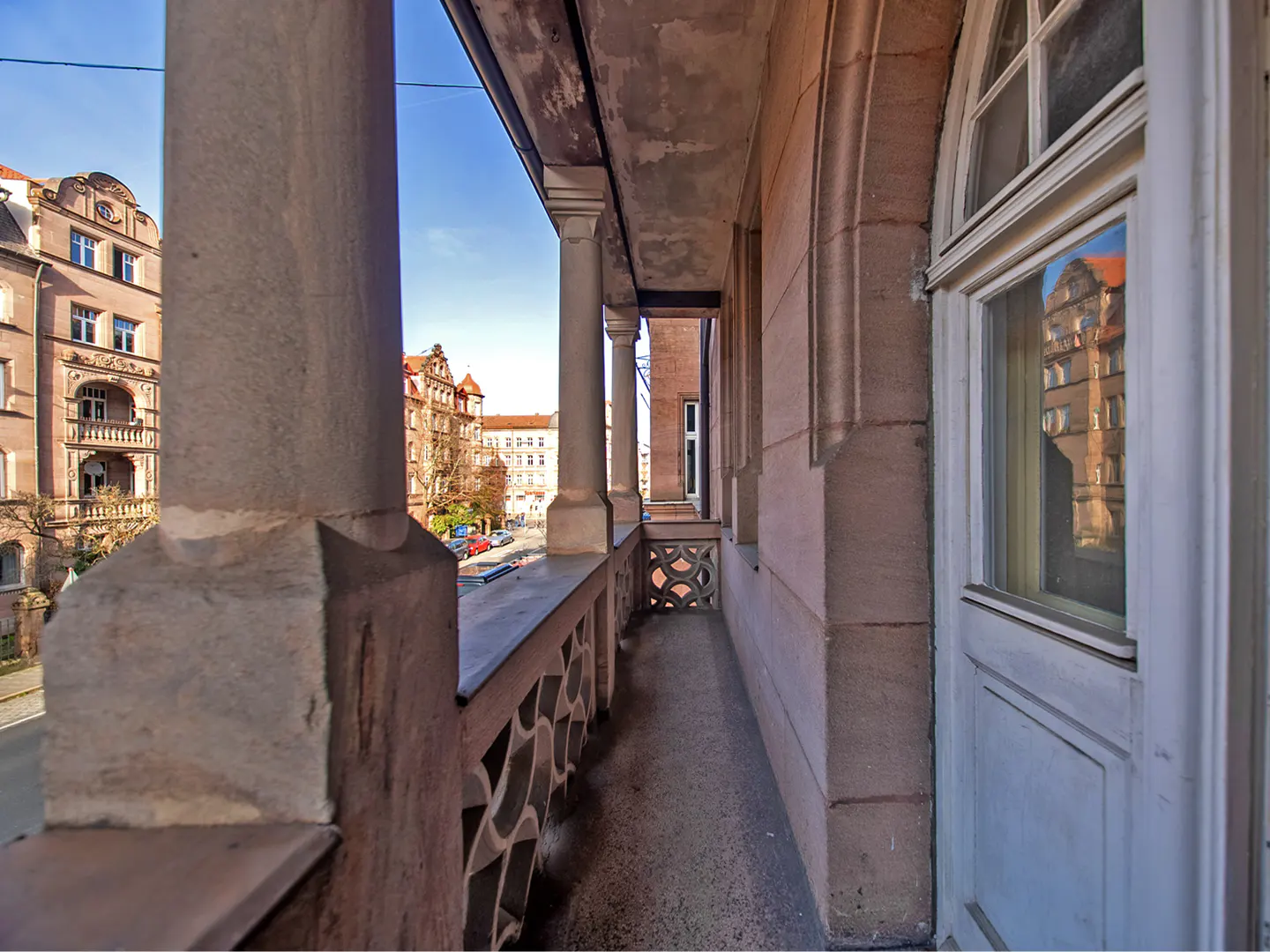 View from a stone balcony with decorative railings and columns, overlooking a European city street with brick buildings.