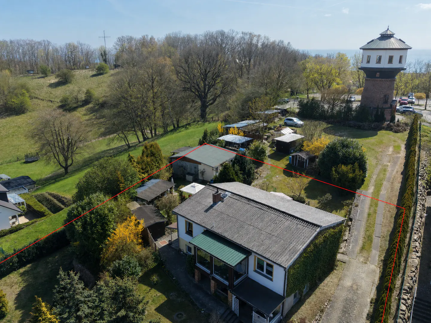 Aerial view of a white two-story house with a gray roof, surrounded by trees and a red property line. A brick water tower is visible in the background.