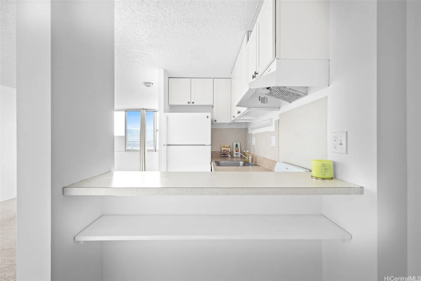 Bright, all-white kitchen with a breakfast bar. Cabinets, fridge, and range hood are visible. A window offers a city view.