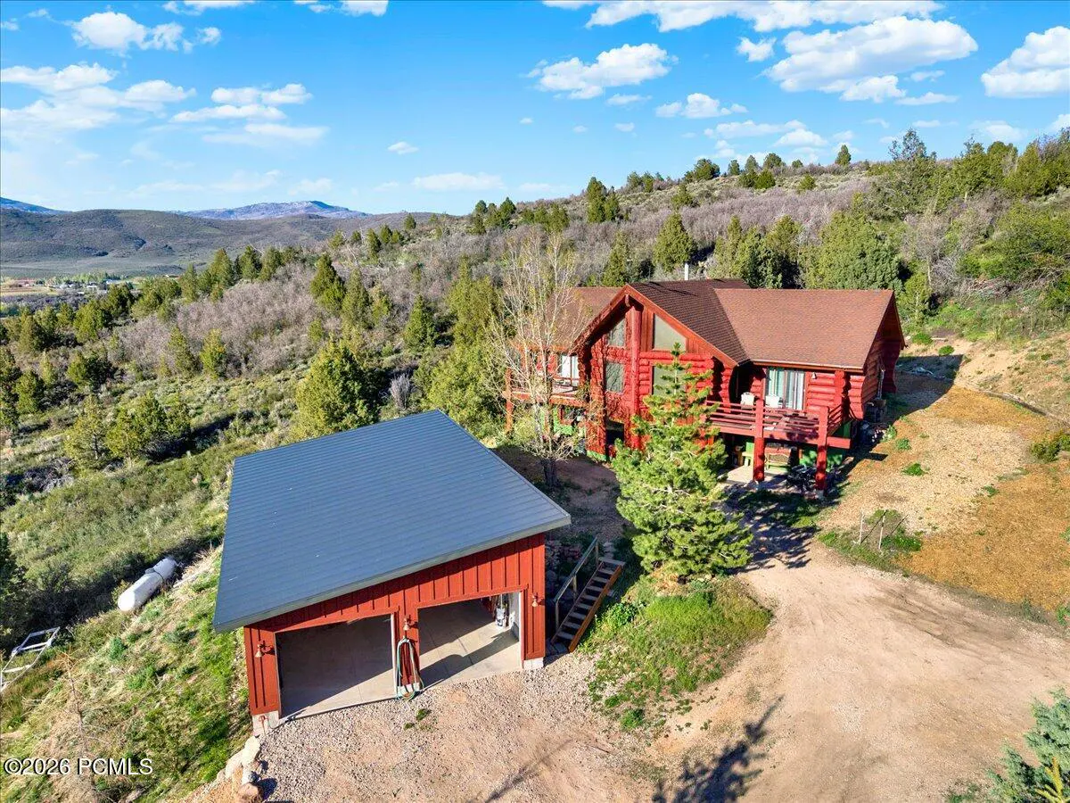 Aerial view of a red log cabin home with a matching garage on a hillside with trees and mountains in the background.