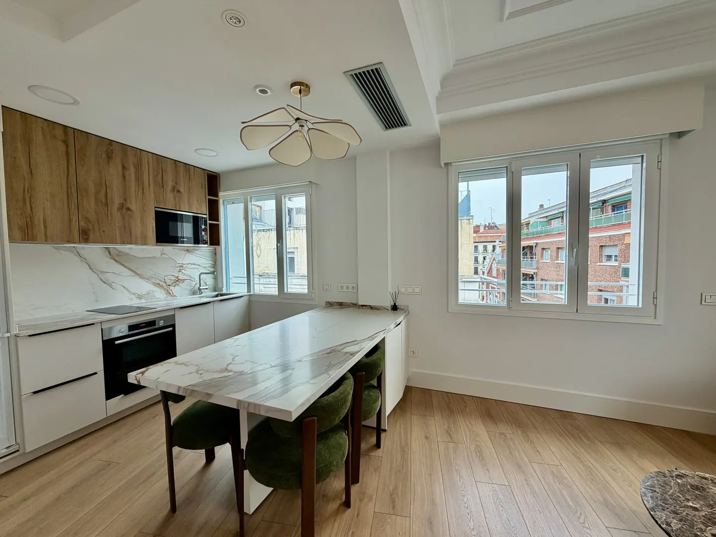 Bright kitchen with wood floors, white cabinets, and marble countertops. A marble island with green chairs sits in the center. Windows overlook a city.