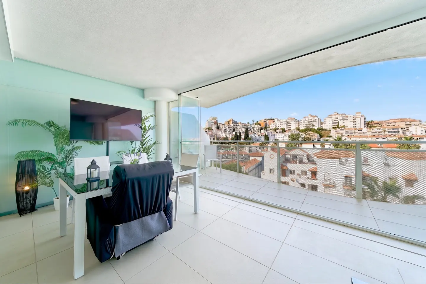 Balcony view of city. Table with black chair cover, TV, and plants inside. Sliding glass doors open to a balcony with a city view.