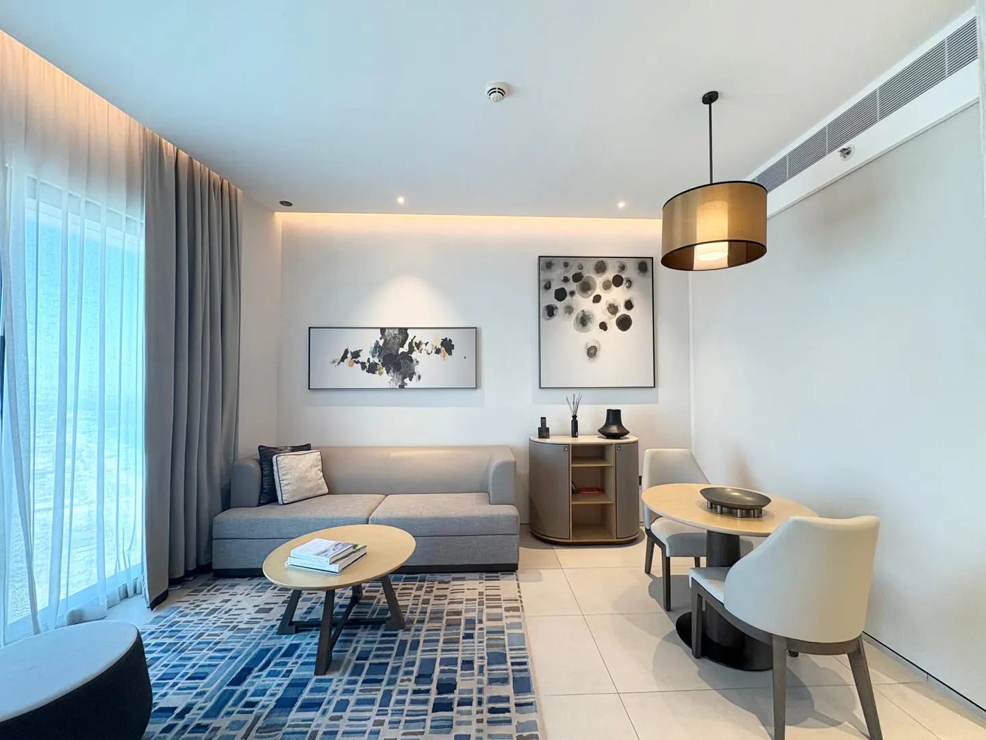 Modern living room with a grey sofa, oval coffee table, blue patterned rug, and a small dining area with two chairs.