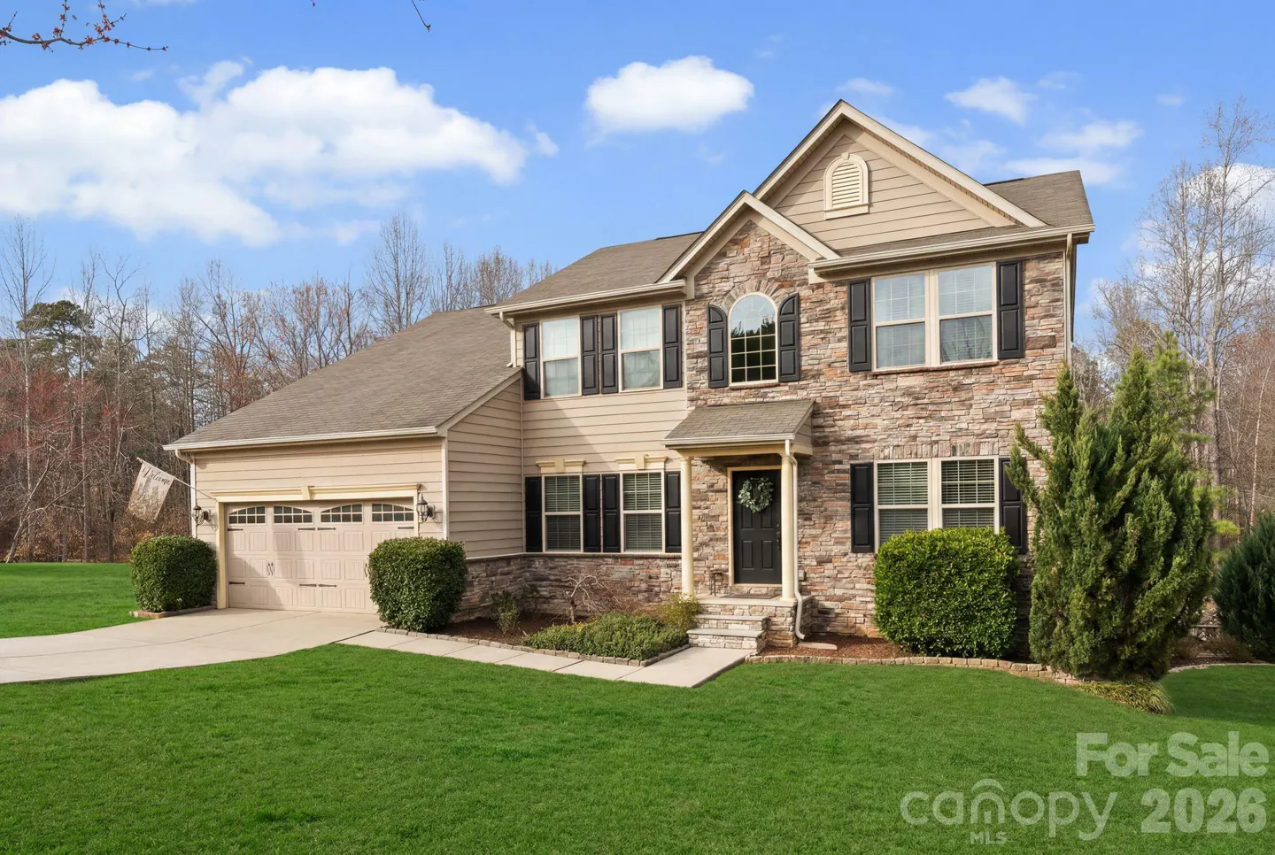 Two-story house with stone and tan siding, black shutters, green lawn, and blue sky.