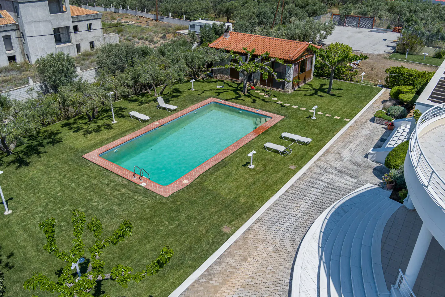 Aerial view of a rectangular pool with turquoise water, surrounded by green grass, lounge chairs, and a small house with a red tile roof.