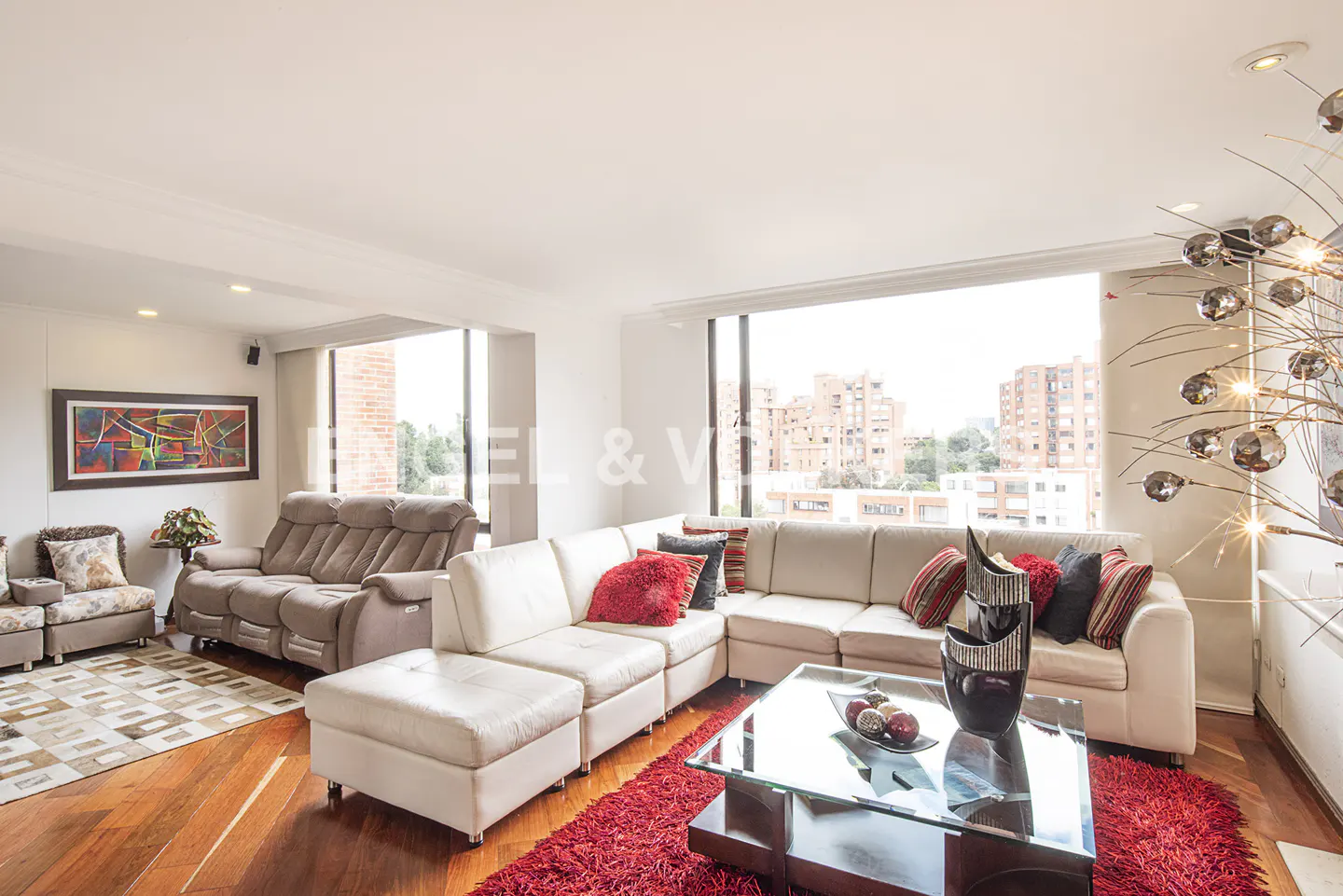 Bright living room with a white sectional sofa, red rug, glass coffee table, and a view of city buildings through windows.