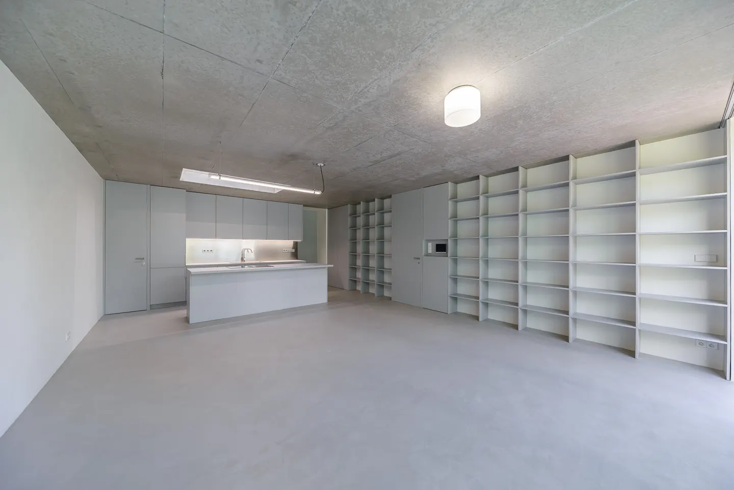 Minimalist kitchen and living space with gray cabinets, island, concrete ceiling, and large empty bookshelf.