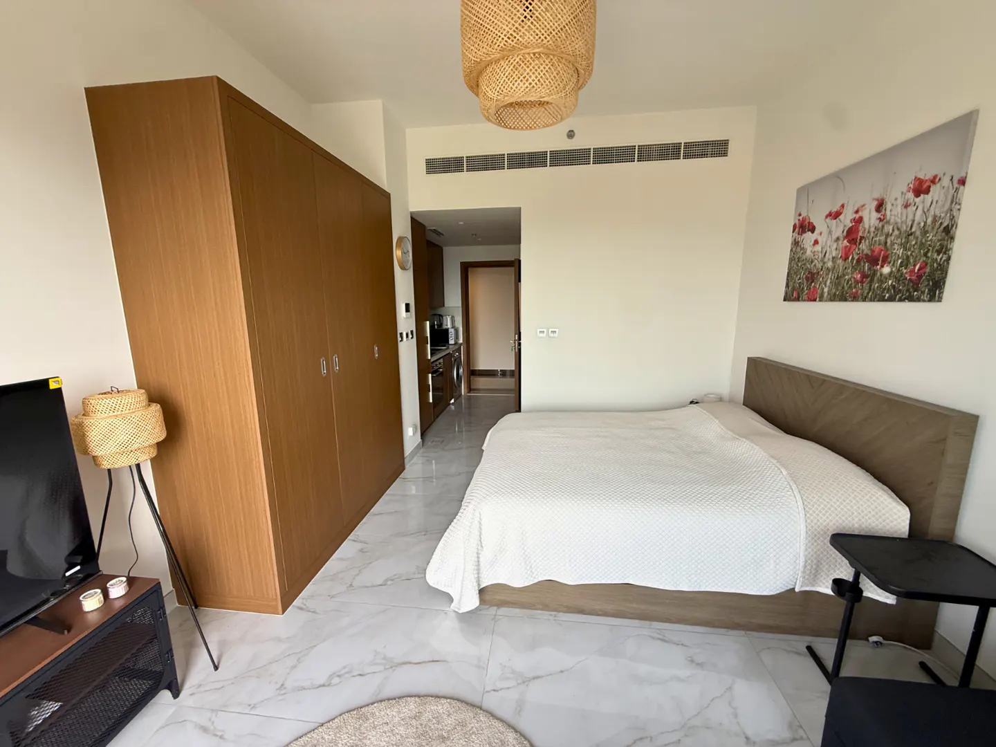 A studio apartment with a bed, wardrobe, TV, and kitchenette. The walls are white, and the floor is marble. A woven lamp hangs from the ceiling.
