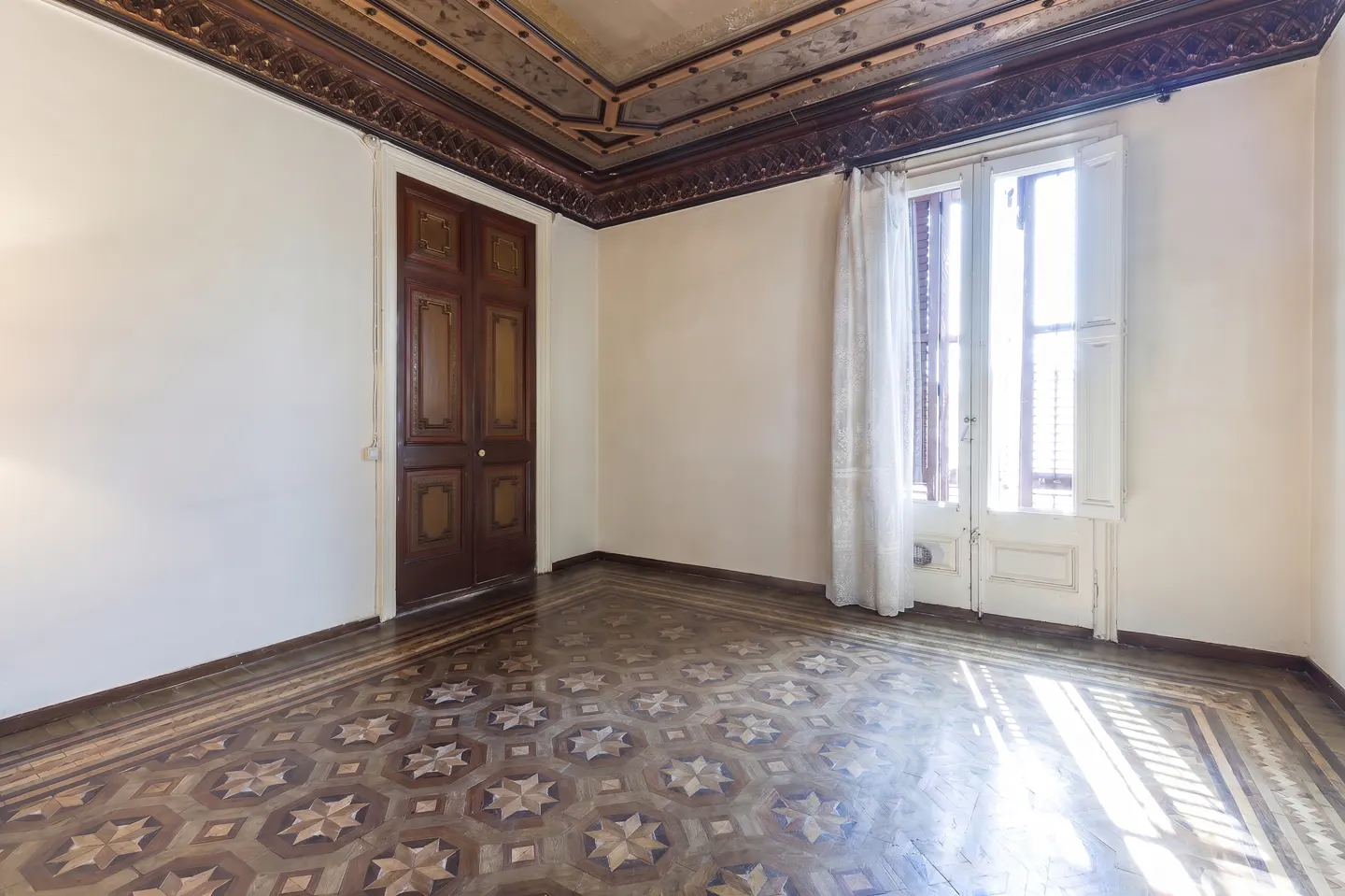 Empty room with decorative ceiling, patterned wood floor, and white walls. A brown door and white window with curtains are visible.