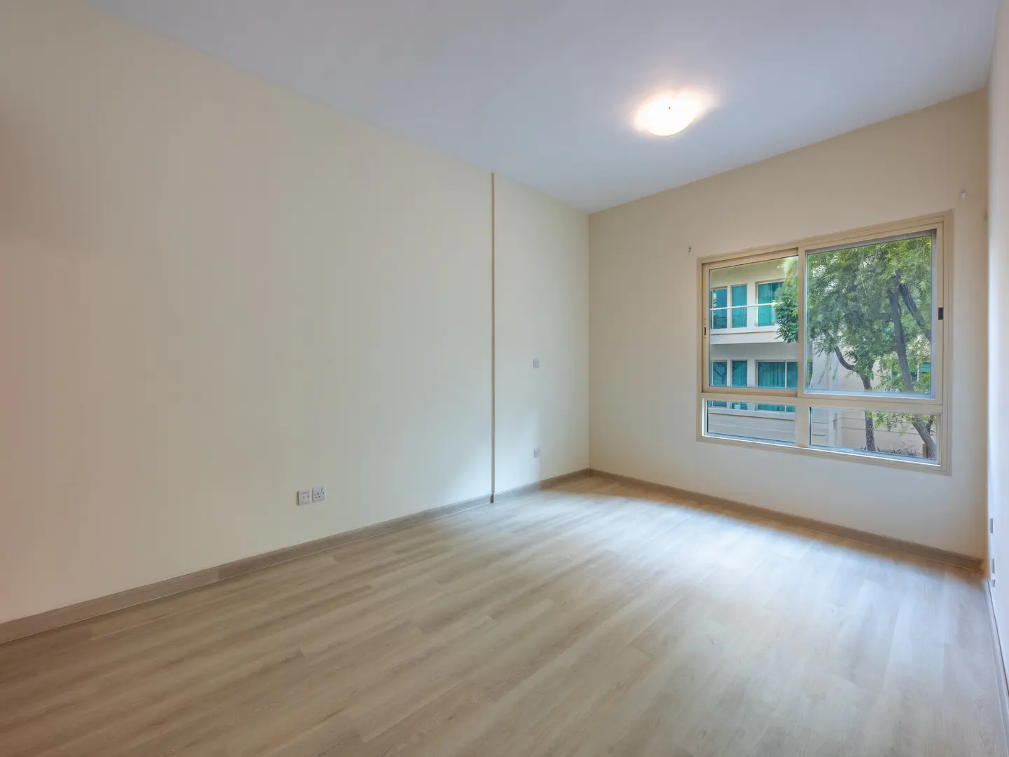 Empty room with light wood floors, cream walls, and a large window. Outside the window are trees and a building.