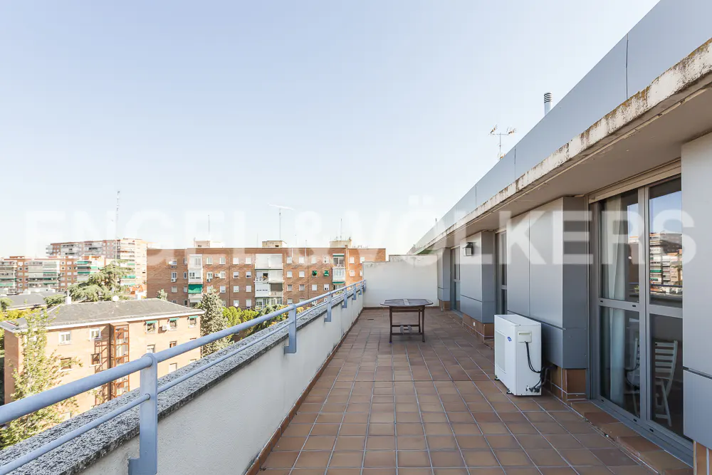 Wide terrace view with brown tile flooring, a small table, and a blue railing overlooking city buildings.