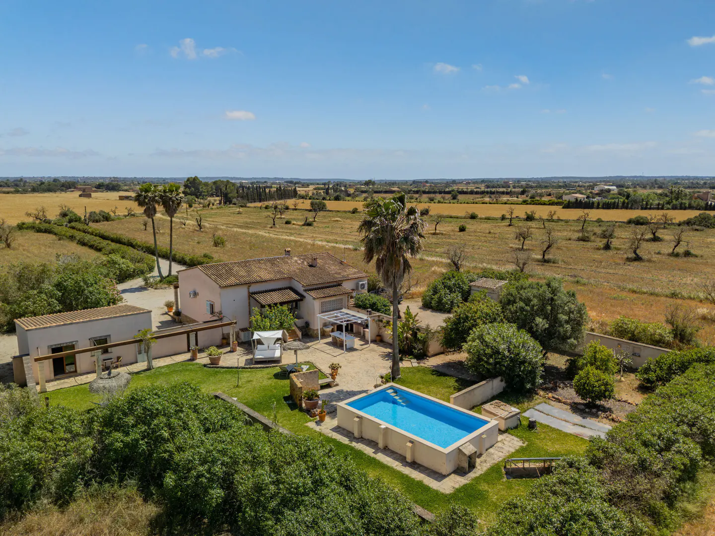 Aerial view of a tan house with a pool, palm trees, and green lawn against a rural backdrop under a blue sky.