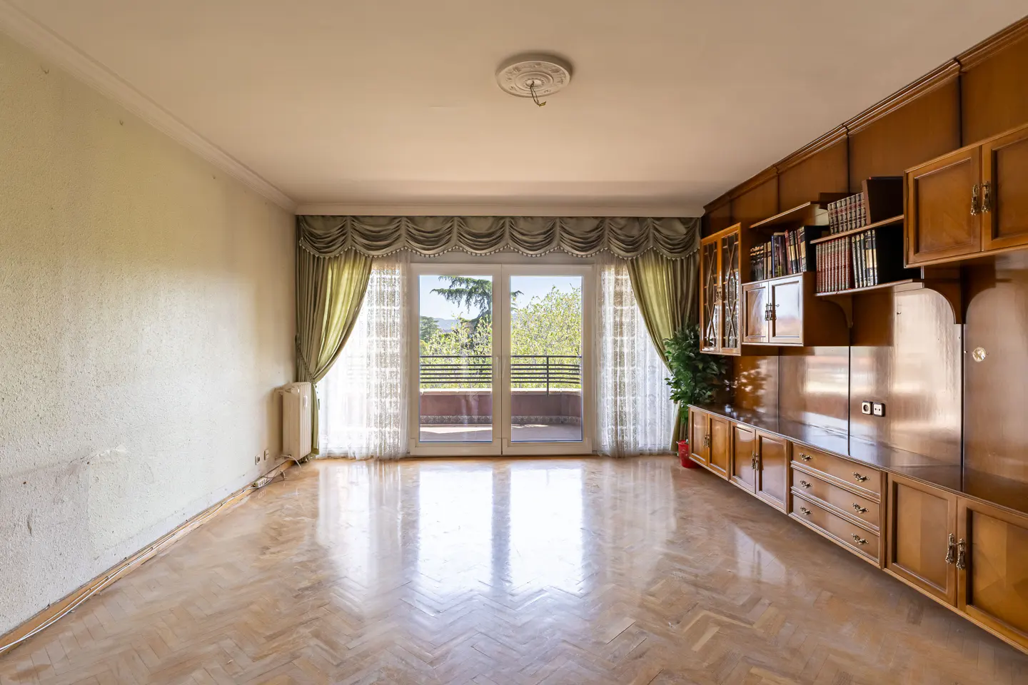 Bright living room with parquet floors, a large window with green curtains, and a built-in wooden cabinet with shelves and drawers.