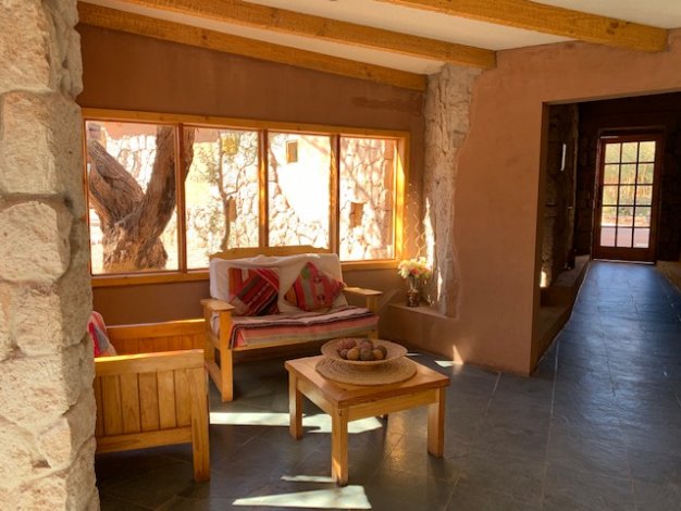 Rustic sunroom with wooden furniture, stone walls, and a view of a tree through a large window. A doorway leads to another room.