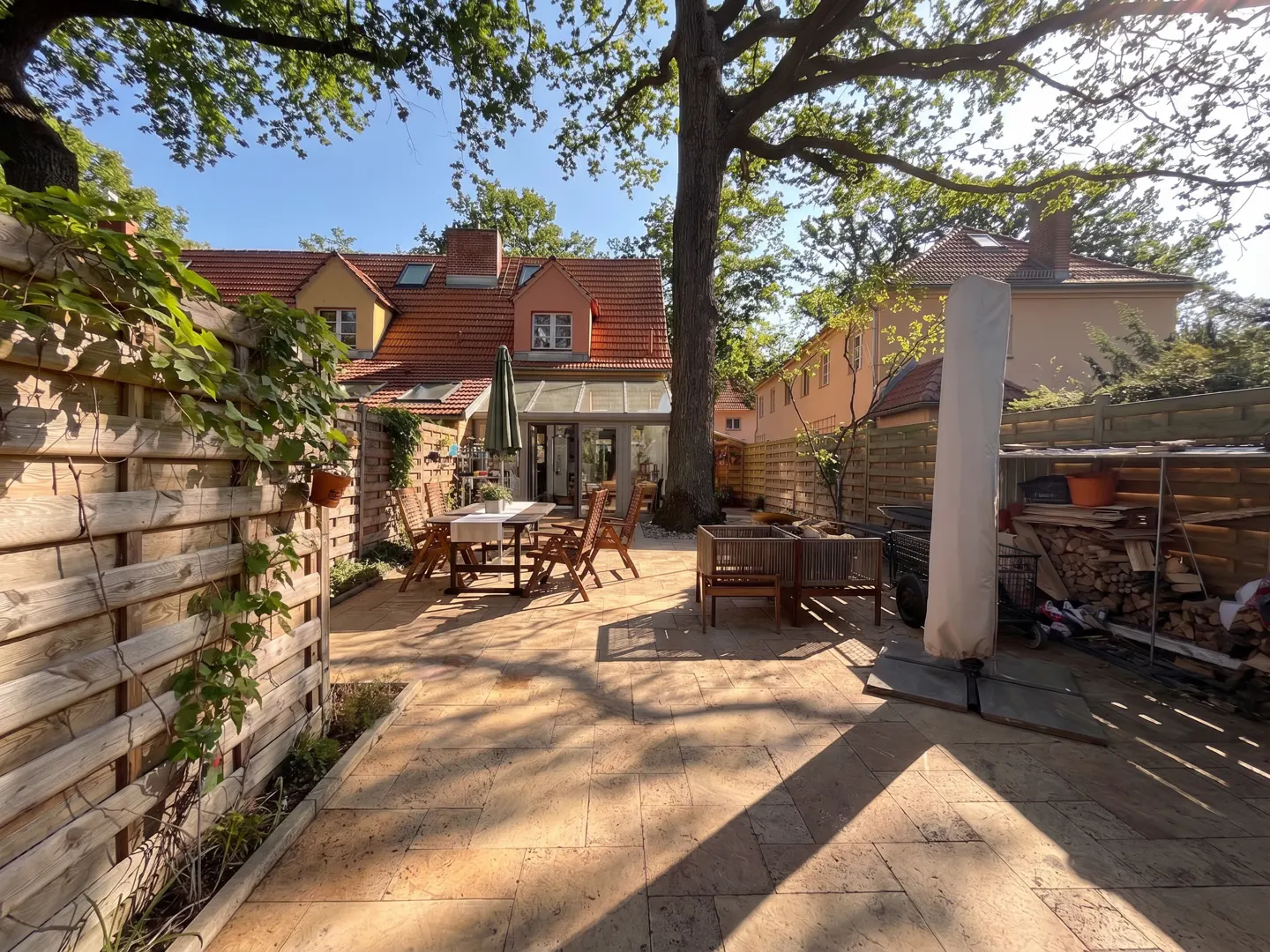 A sunny backyard patio with a dining table, wooden fence, and a large tree providing shade. A woodpile is visible in the background.