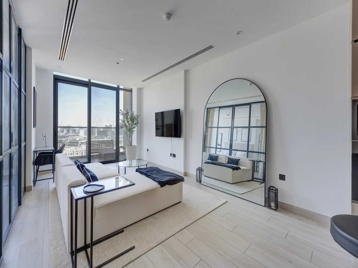 Bright living room with white walls, light wood floors, and a large arched mirror reflecting the space. A white sofa faces a balcony with city views.