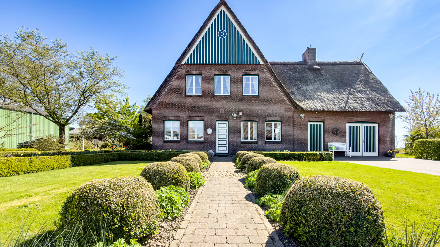 A brick house with a thatched roof, green accents, and a stone path leading through a manicured garden under a blue sky.