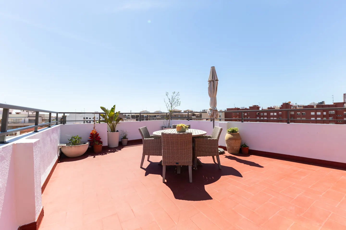 Rooftop patio with a round table, four chairs, and a closed umbrella. Potted plants line the white walls, and a metal railing surrounds the space.