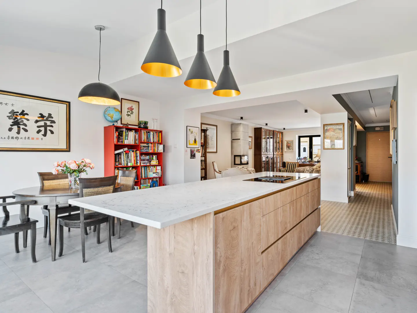 Open-concept kitchen with a large island, dining table, and red bookshelf. Three pendant lights hang above the island.