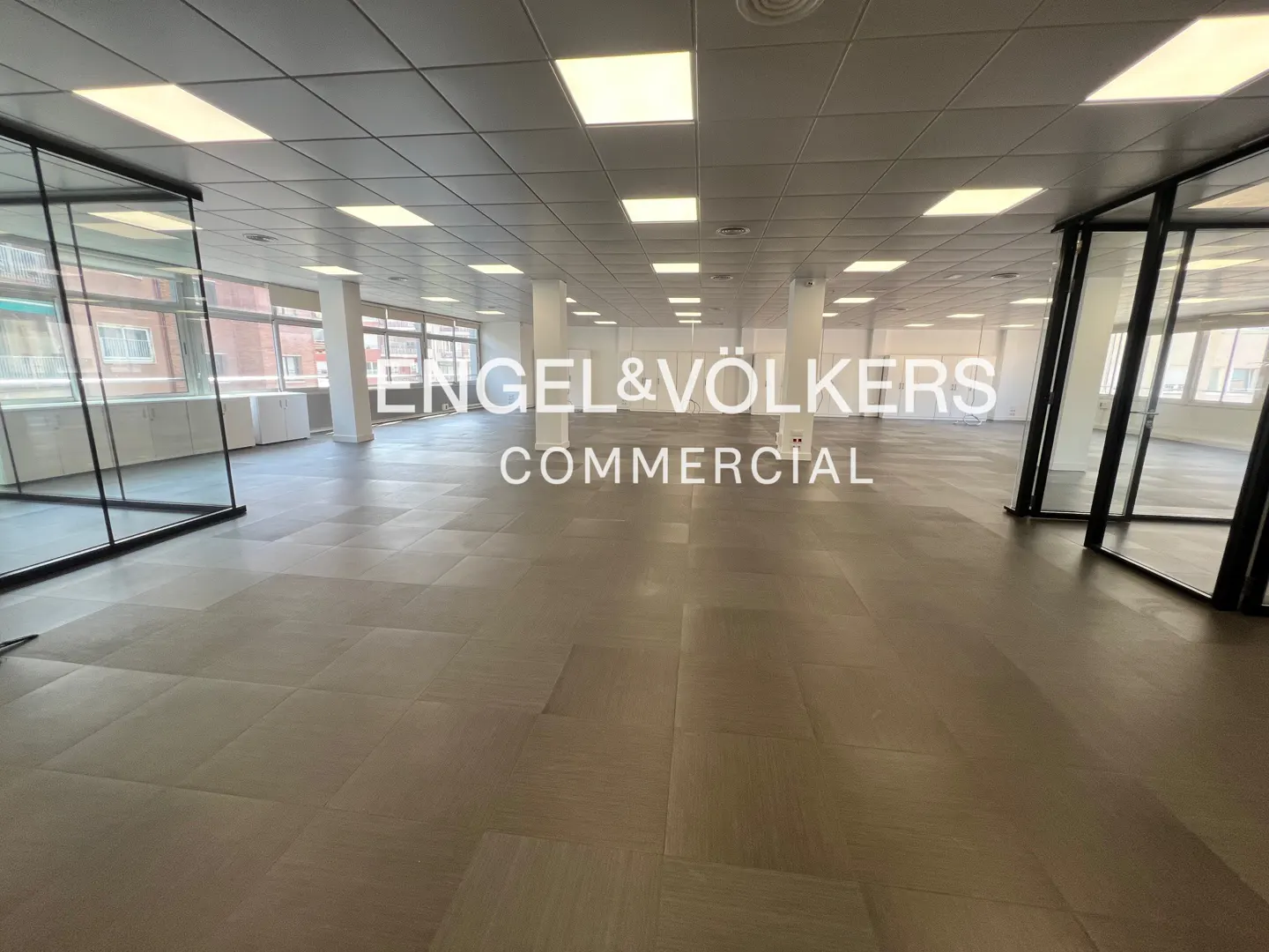 Bright, empty office space with gray floors, white walls, and large windows. Glass-walled rooms on either side. "Engel & Völkers Commercial" overlay.