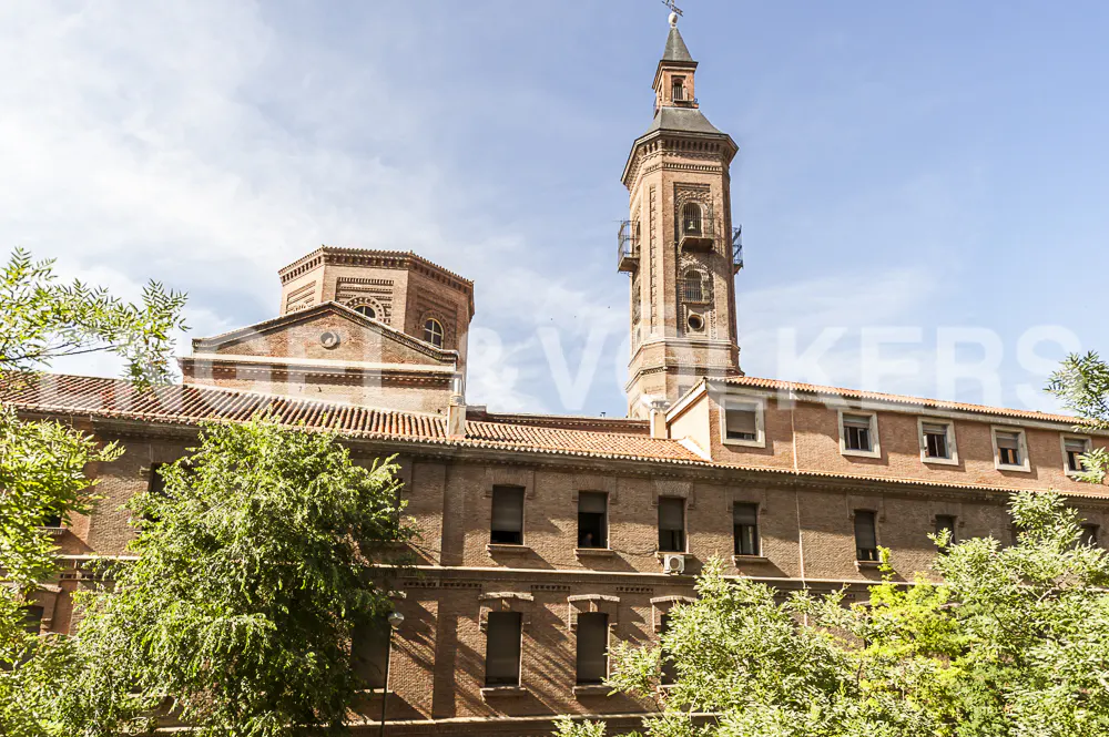 Exterior view of a brick building with a tall tower, red tile roof, and green trees in the foreground.