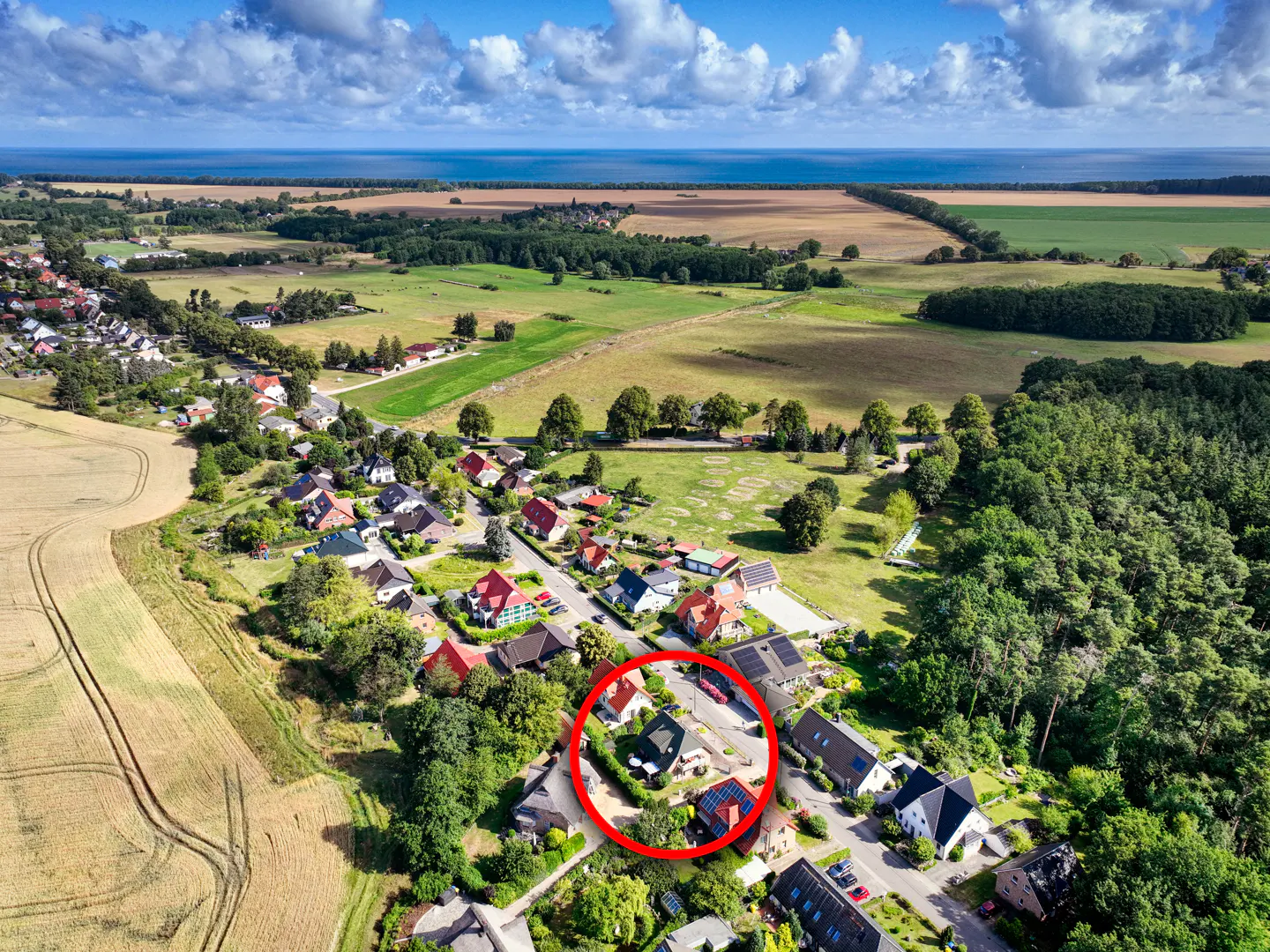 Aerial view of a village with fields, forest, and the sea in the background. A red circle highlights a property.