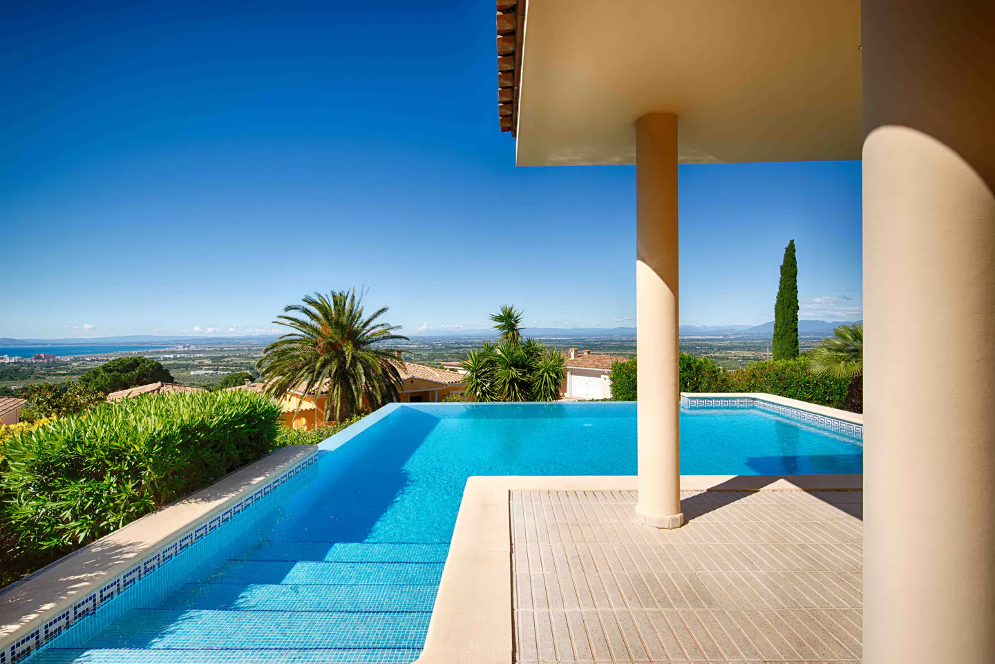 Infinity pool with blue tiles and beige patio, overlooking a green landscape and blue sky. Two beige pillars are visible.
