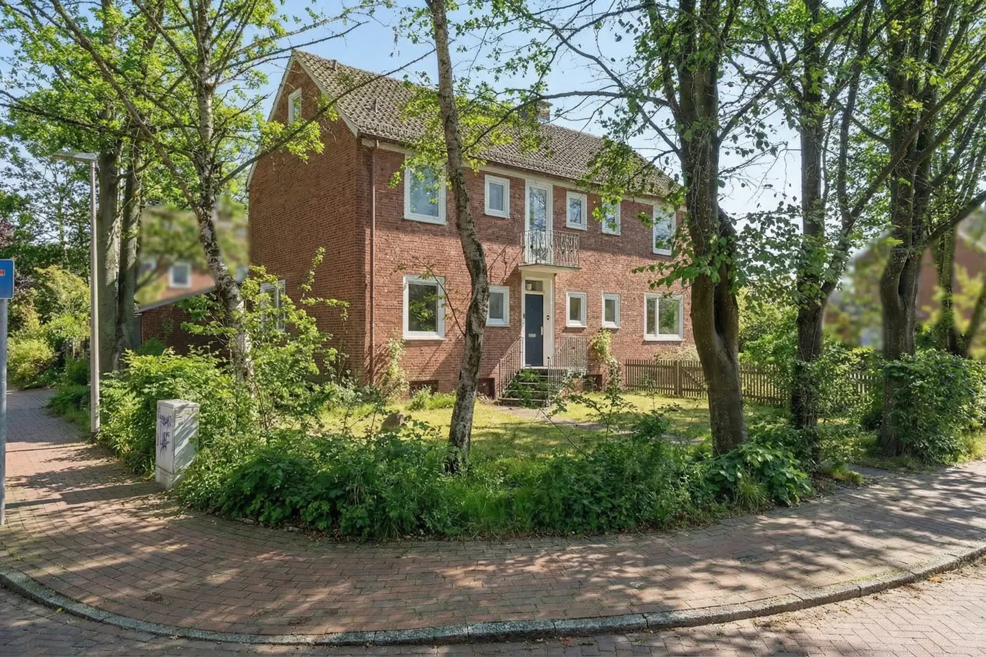 A two-story brick house with a dark blue door and white trim, surrounded by trees and greenery.