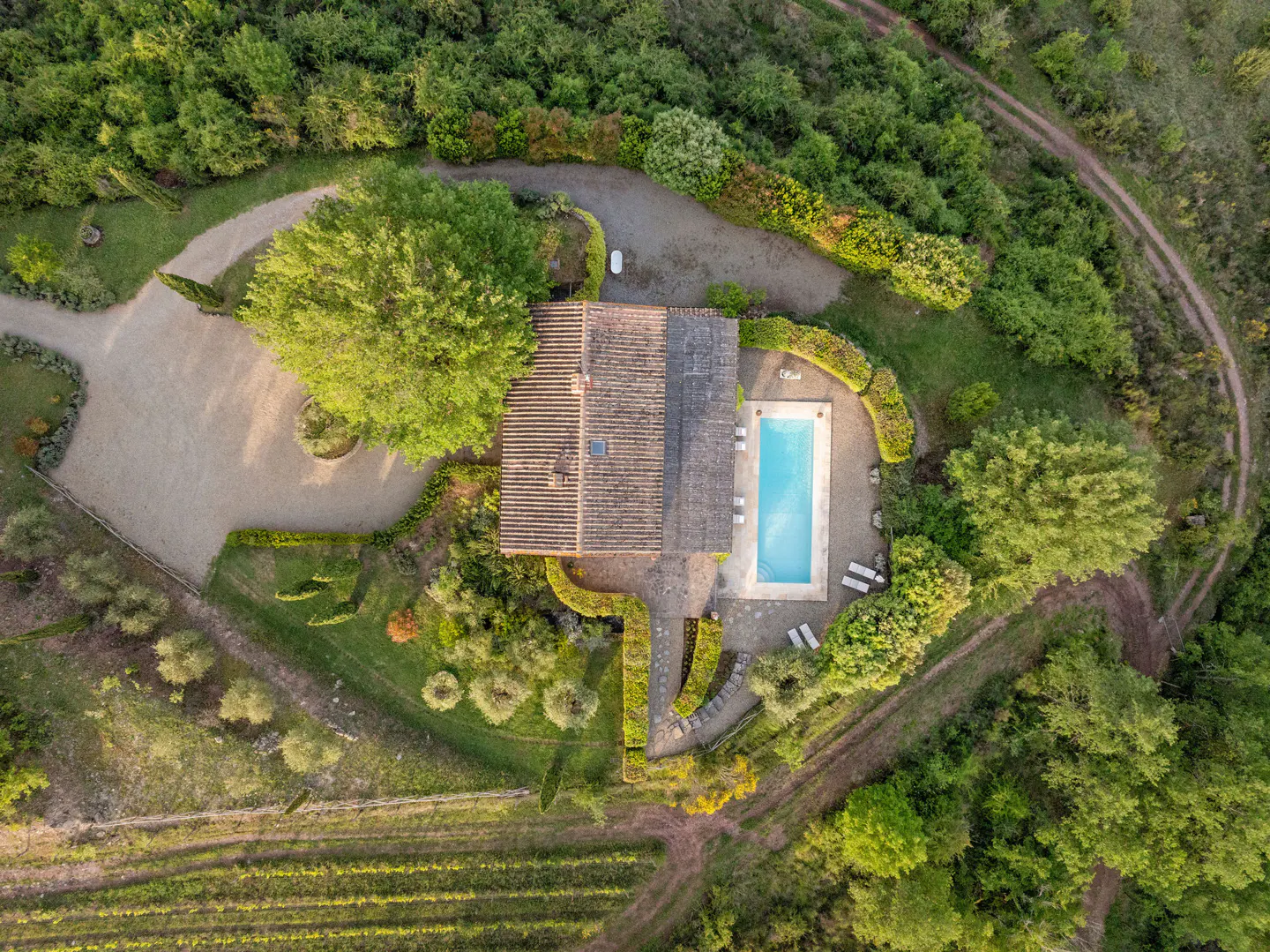 Aerial view of a Tuscan villa with a pool, surrounded by lush greenery and vineyards. The villa has a terracotta roof and a gravel driveway.