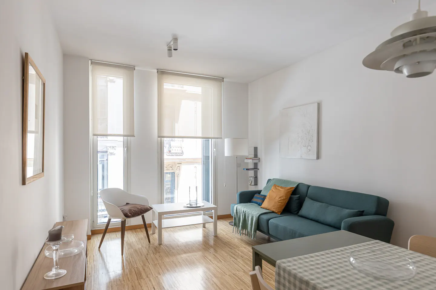 Bright living room with wood floors, white walls, and large windows. A teal sofa with pillows sits near a white coffee table and chair.