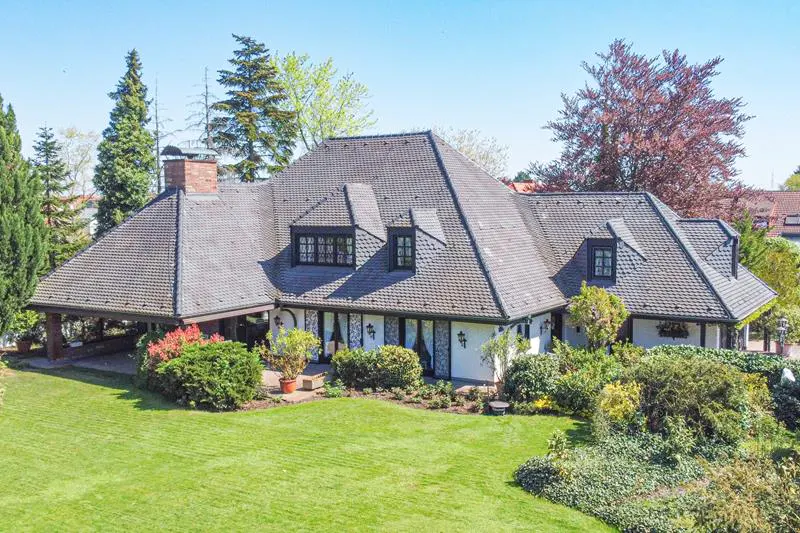 A large, white Tudor-style house with a gray tile roof sits on a green lawn surrounded by trees and bushes.