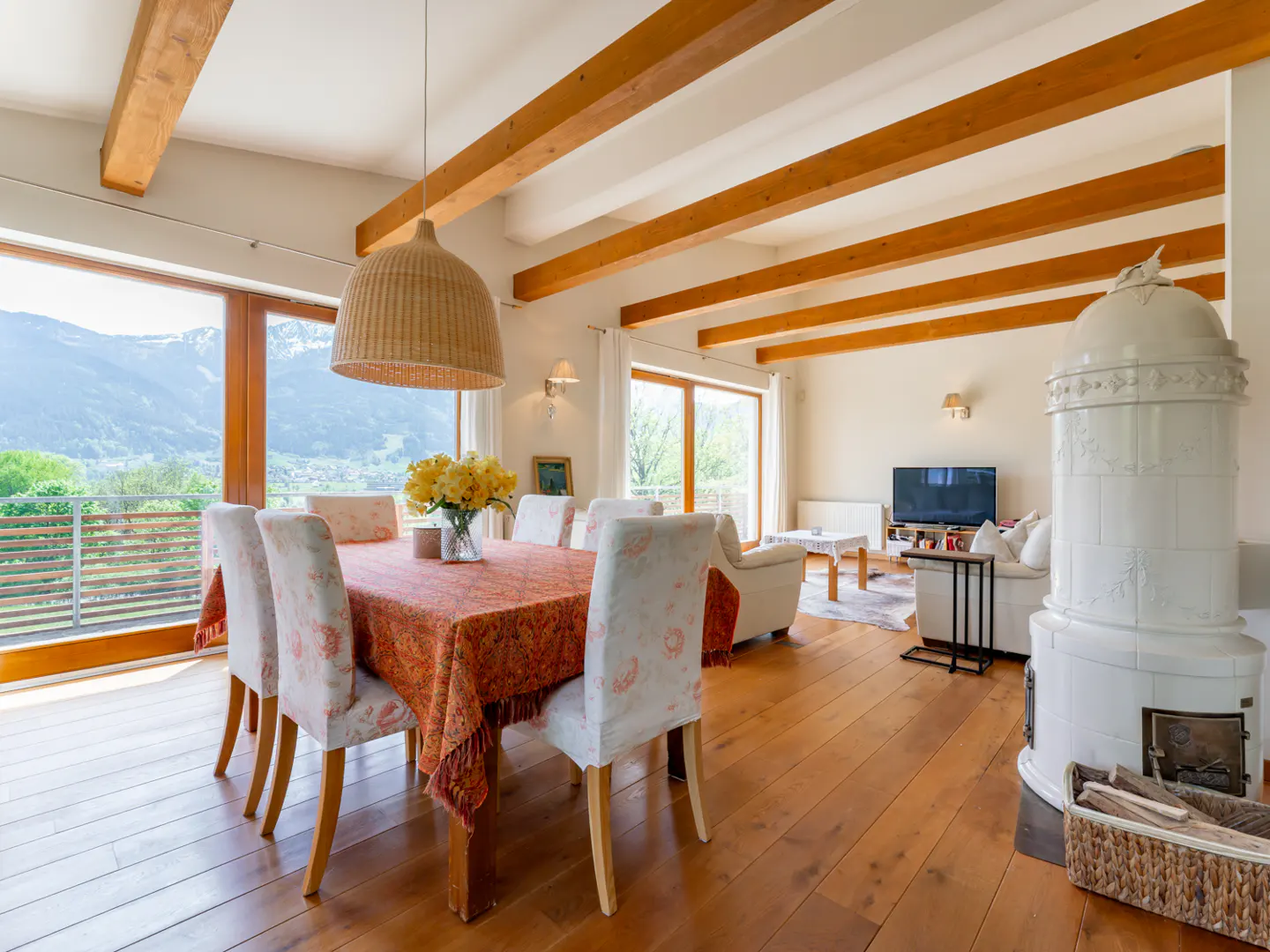 Bright, open-plan living and dining area with wood floors, exposed beams, and mountain views. Table set with floral tablecloth and yellow flowers.