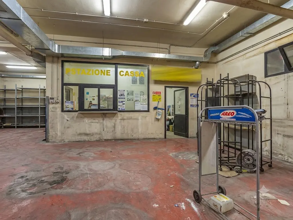 Industrial warehouse interior with a red concrete floor, metal shelving, and a cashier office window labeled "CASSA".
